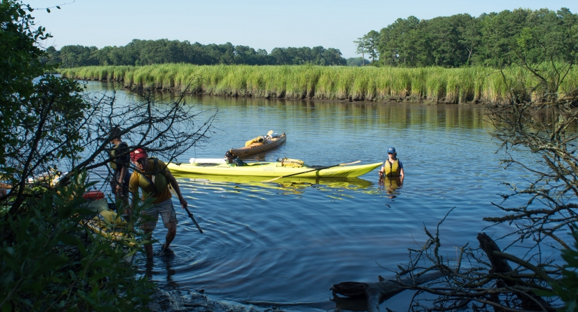 a person stands in waist-deep water beside a couple of floating kayaks, while others navigate a different kayak toward the shore. 